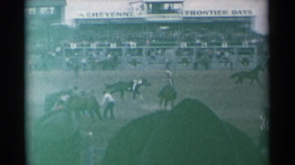 Wide 8mm view of the Cheyenne Frontier Days rodeo arena with horses spread across the field, cowboys on foot, spectators, and foreground silhouettes.