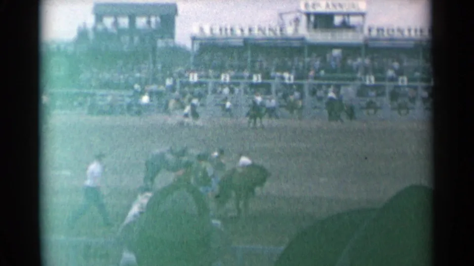 Wide 8mm view of the Cheyenne rodeo arena, showing horses and participants below a crowded grandstand and announcer structure.