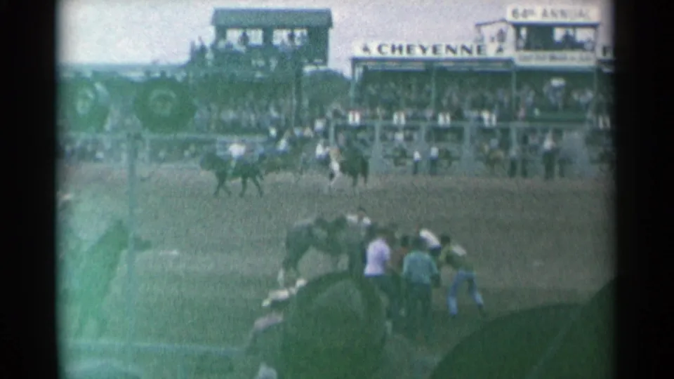 Home-movie view of the Cheyenne rodeo arena with mounted riders, nearby horses and spectators in the stands.