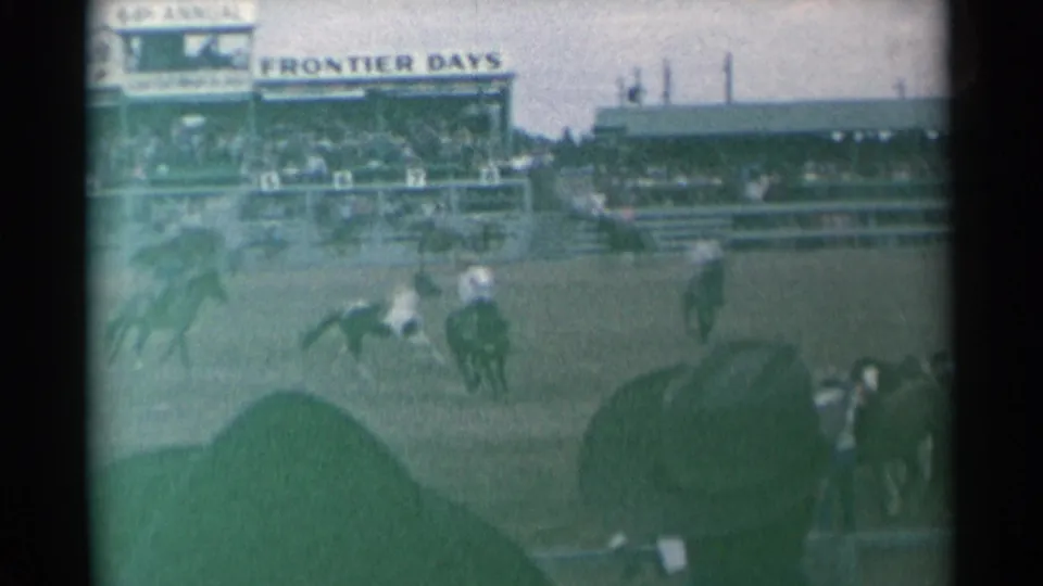 Wide 8mm frame of riders moving across the Cheyenne rodeo arena with grandstands and Frontier Days signage visible.