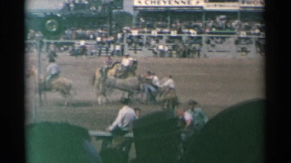 Arena hands and mounted riders cluster around horses at the 1961 Cheyenne rodeo, with the grandstand crowd and signage behind them.
