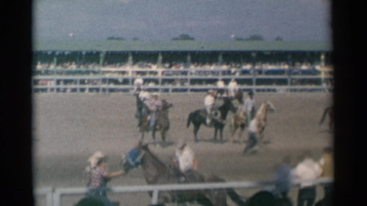Bucking horse and rider in a rodeo arena with mounted cowboys nearby and spectators in bleachers.