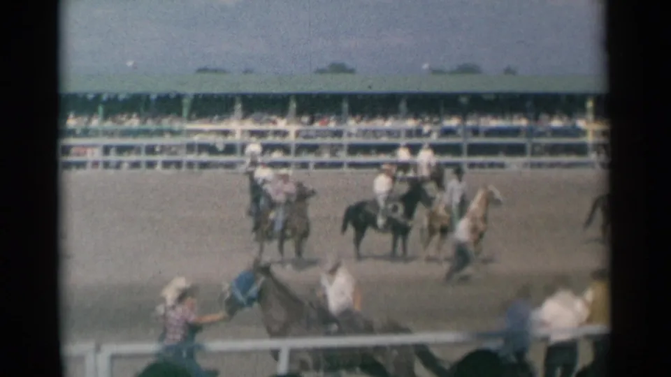 1961 Cheyenne rodeo view of a bronco ride in a dirt arena, with mounted cowboys, railings, and spectators visible beyond the action.