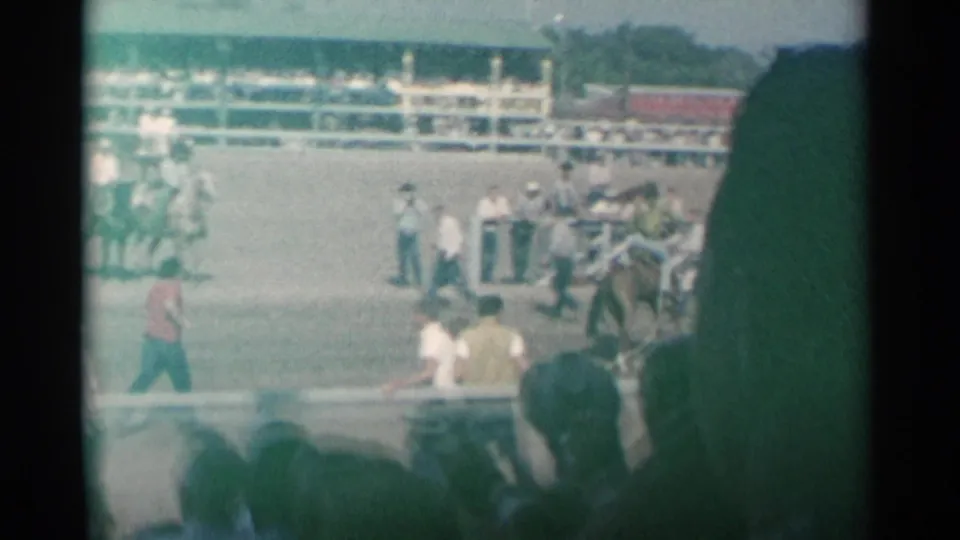 Home-movie view across a Cheyenne rodeo arena, showing a horse near the rail, rodeo personnel, mounted riders, and a crowd in the stands.