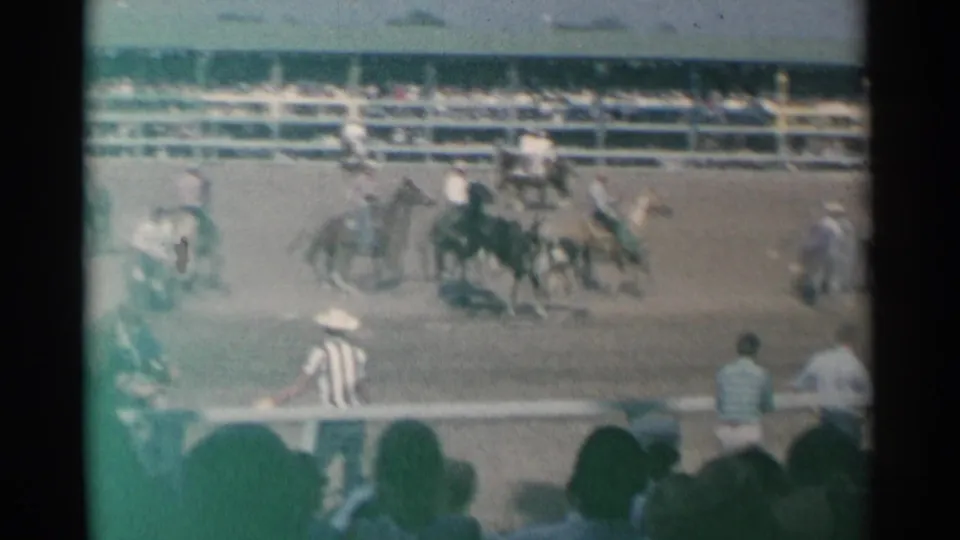 Cheyenne rodeo arena after the main action, with mounted cowboys, arena staff, railings, bleachers, and foreground spectators in soft 8mm color.