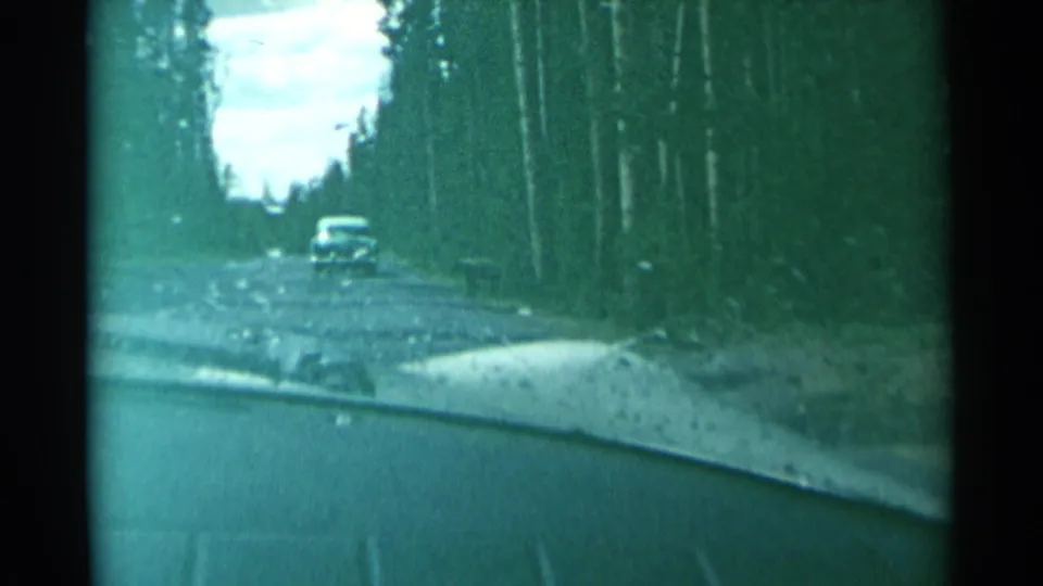 View from inside a moving car on a narrow wooded road, with a distant automobile ahead in 1961 Wyoming footage.