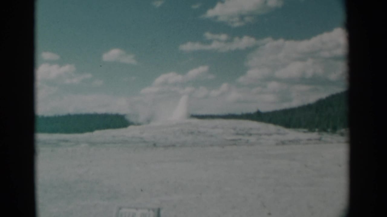 Distant geyser plume rising from a pale basin under scattered clouds.