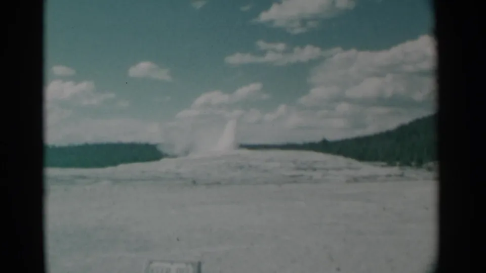 1961 8mm home-movie frame showing a distant geyser eruption across an open basin, with forested hills and a partly cloudy sky.