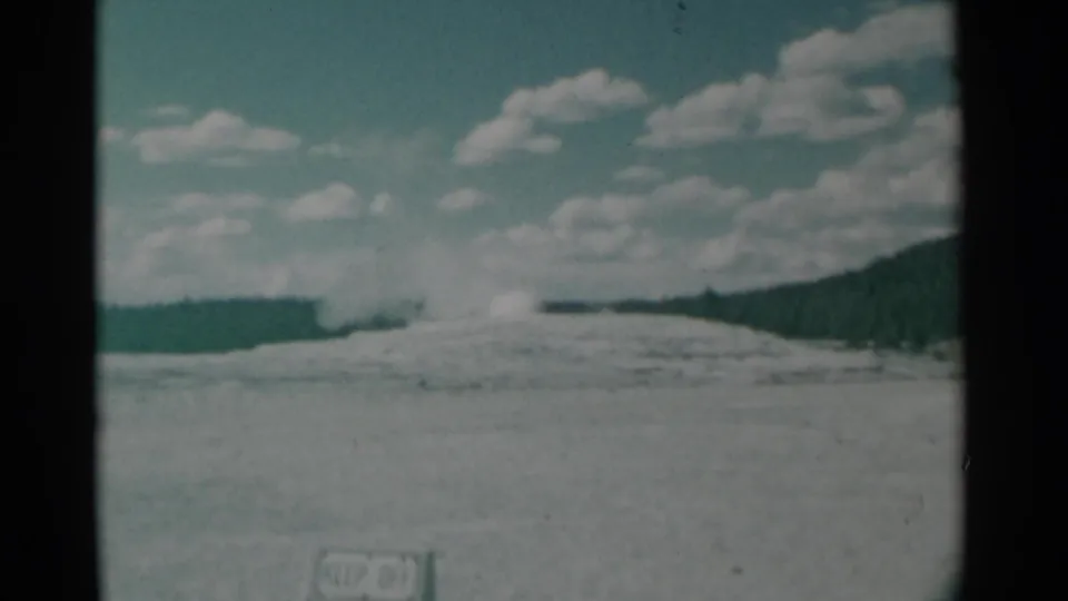 Soft-focus 1961 8mm frame of a geyser eruption seen from across an open basin, with hills and scattered clouds beyond.