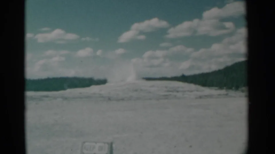 Final 1961 8mm frame shows the geyser plume tapering above the pale basin, with a dark forested slope and clouds behind it.