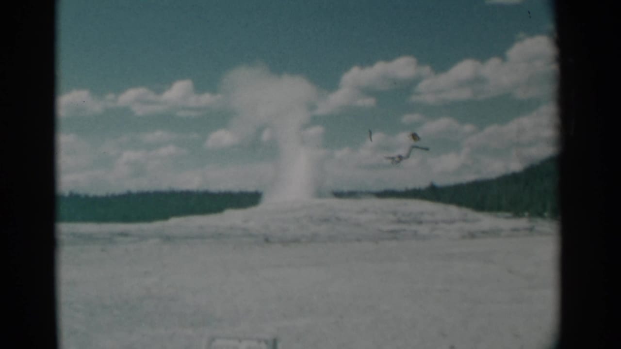 1961 8mm home-movie frame showing a geyser plume rising from a pale mound with distant trees and clouds in the sky.