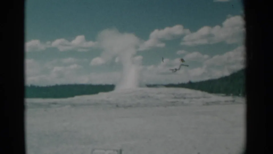 1961 8mm home-movie frame showing a geyser plume rising from a pale mound with distant trees and clouds in the sky.