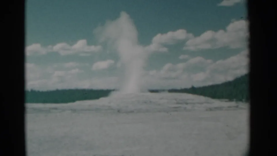 1961 8mm frame of a tall geyser and steam plume rising above a pale basin with distant trees and a cloudy blue sky.