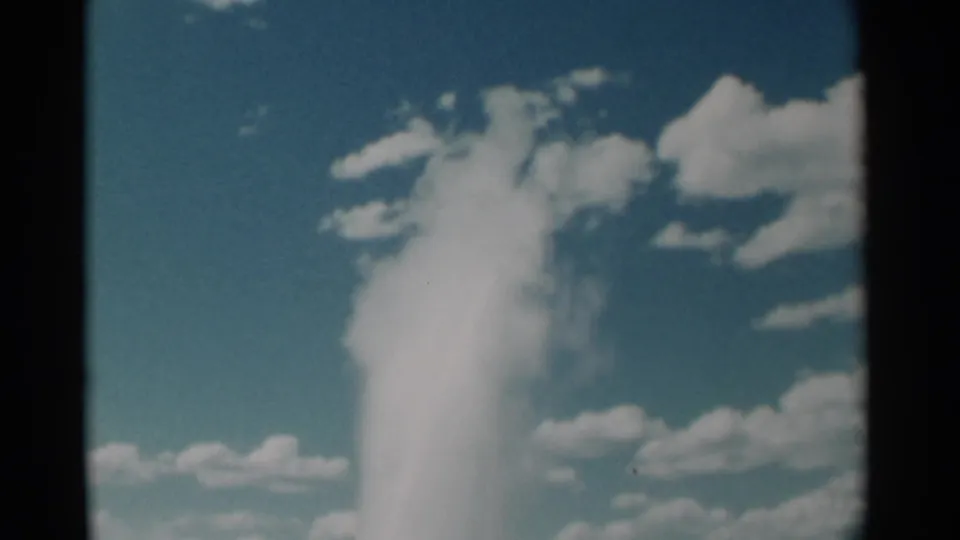 1961 8mm frame showing the geyser plume rising and dispersing into a cloudy blue sky, bordered by dark film edges.