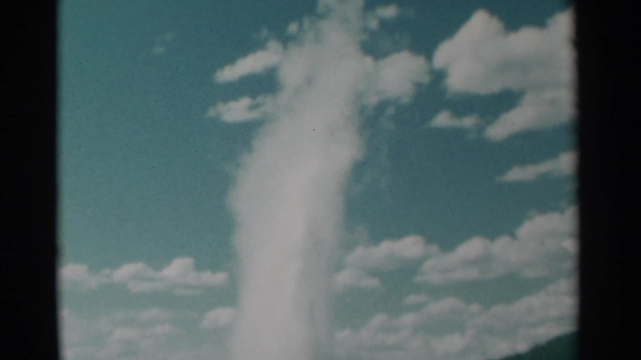 Tall geyser plume rising against a teal sky with clouds and low hills.