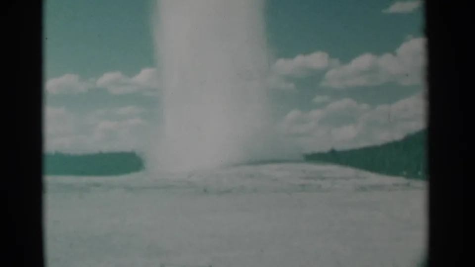 1961 8mm frame of a broad white geyser burst rising from a pale basin, with low hills and scattered clouds behind it.