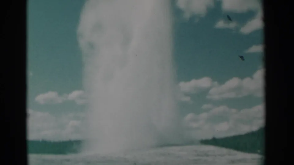 1961 8mm frame with a large geyser plume dominating the right side, set against clouds, teal sky, and a low dark hillside.