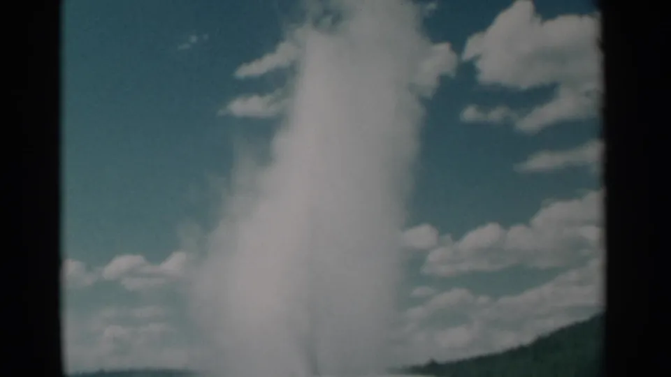 1961 8mm Wyoming footage shows a geyser plume shooting upward against blue sky, clouds, and a low hillside.