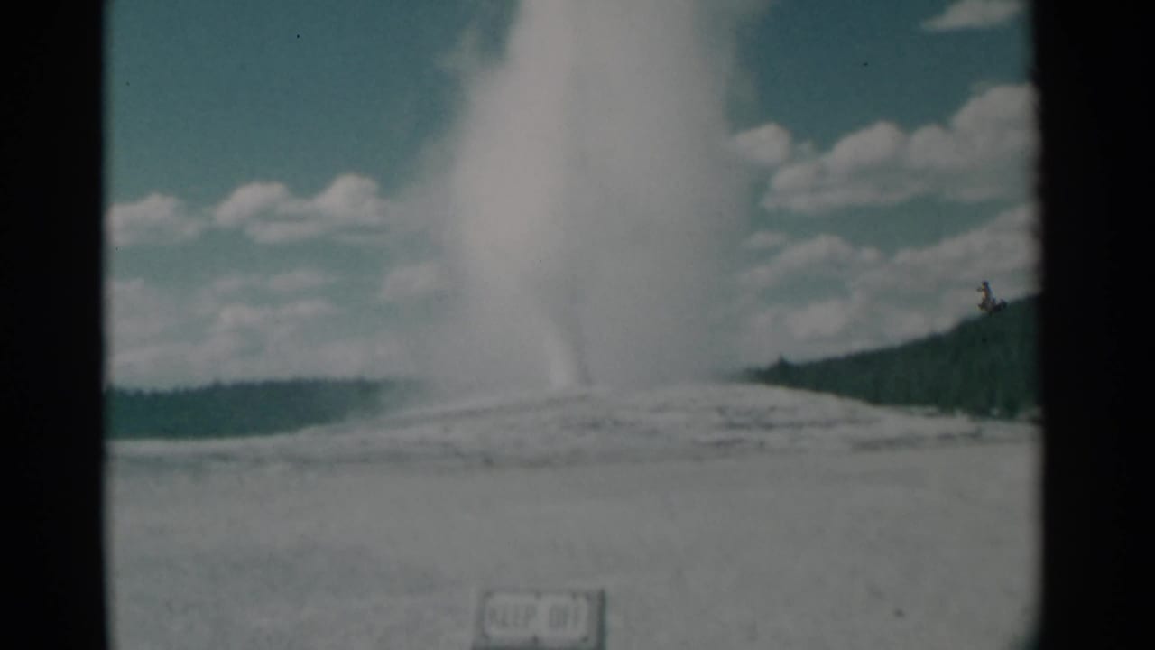 Wide view of a geyser plume rising from open ground below a cloudy blue sky.