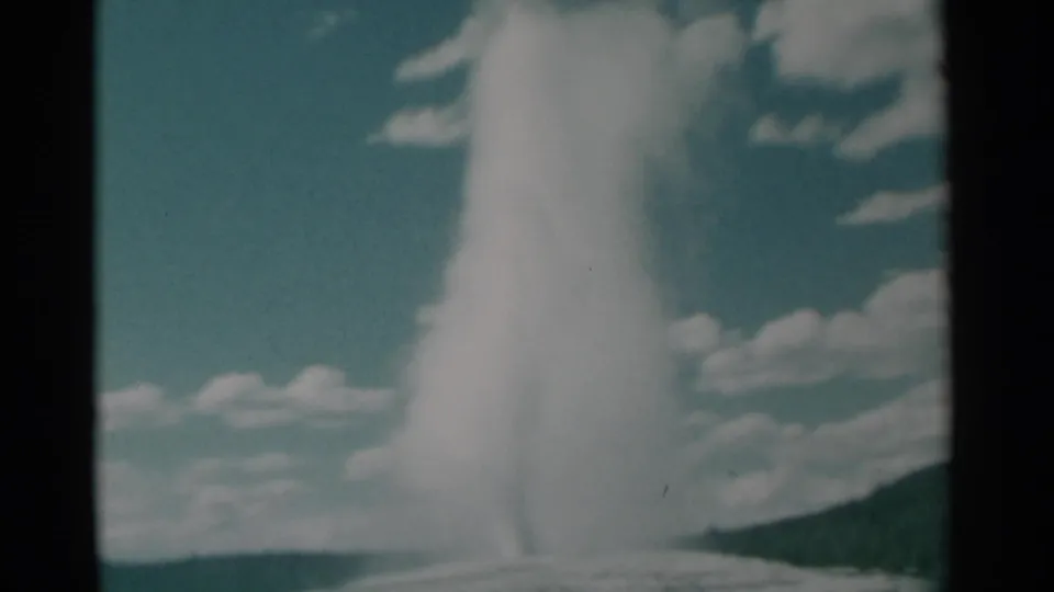 A bright water-and-steam column fills much of the 8mm frame, with blue sky, clouds, and a low Wyoming hillside behind it.