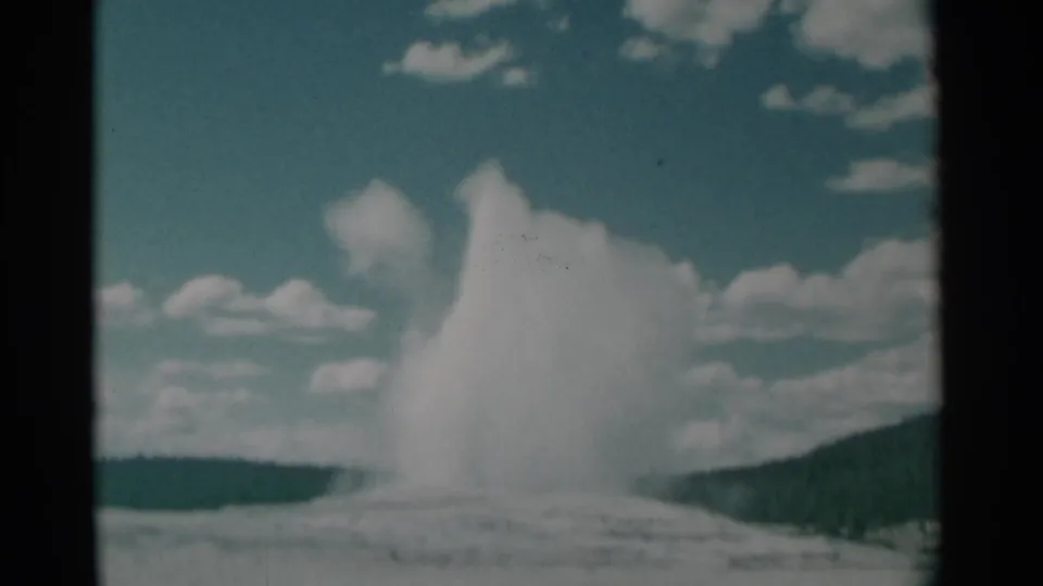 1961 Wyoming 8mm frame showing a geyser erupting from a pale basin, with clouds and a dark ridge in the background.