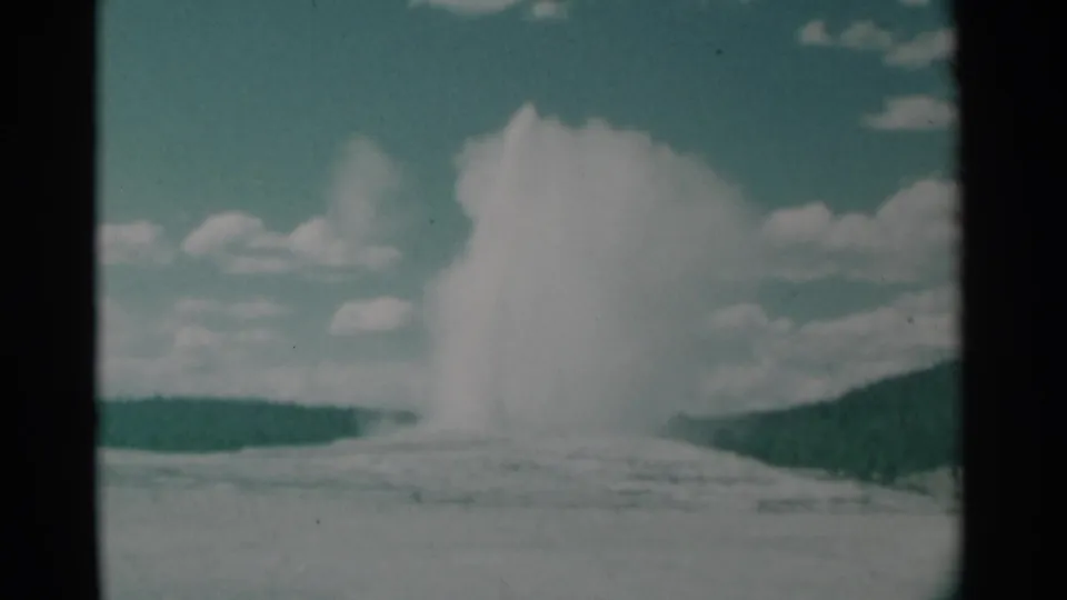 1961 Wyoming 8mm frame showing a broad geyser plume over a pale geothermal basin, backed by a dark ridge and clouded sky.