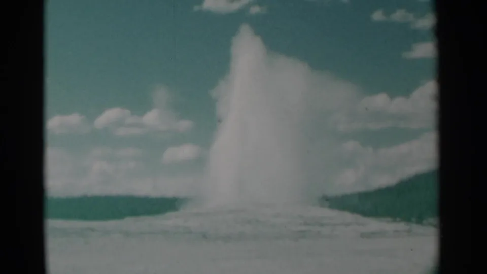 1961 8mm home-movie frame of Old Faithful erupting in Yellowstone, with steam rising over the pale geyser basin and distant tree line.