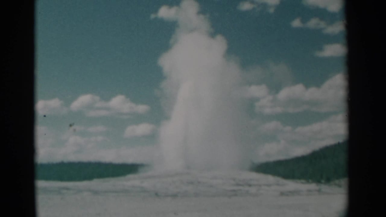 Tall geyser plume rising from a pale basin with clouds and hills behind it.