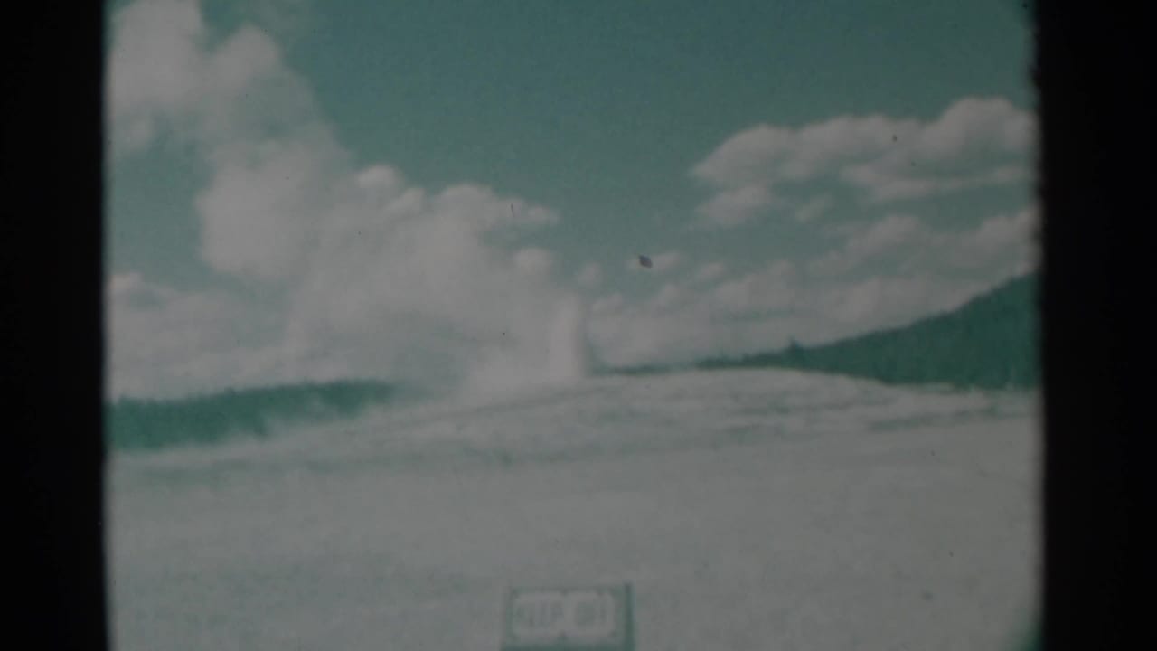 1961 Wyoming 8mm frame showing a distant geyser-like steam plume on a low hill under a blue-green sky with clouds and a small foreground sign.