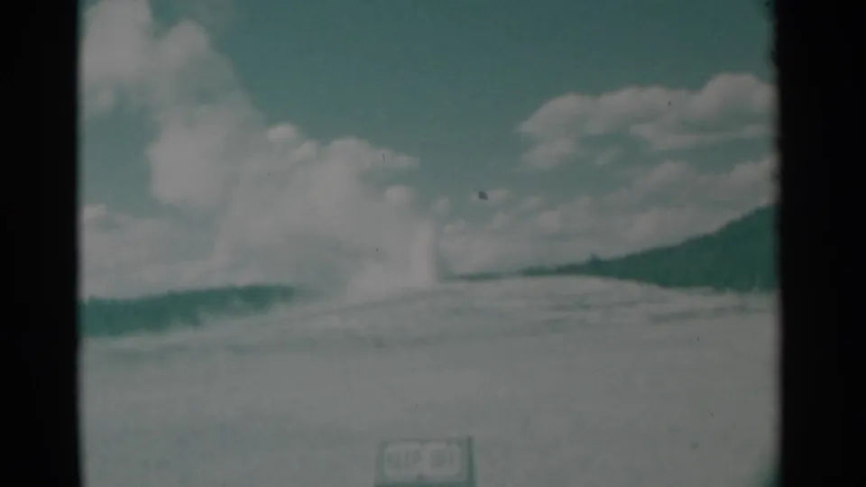 1961 Wyoming 8mm frame showing a distant geyser-like steam plume on a low hill under a blue-green sky with clouds and a small foreground sign.
