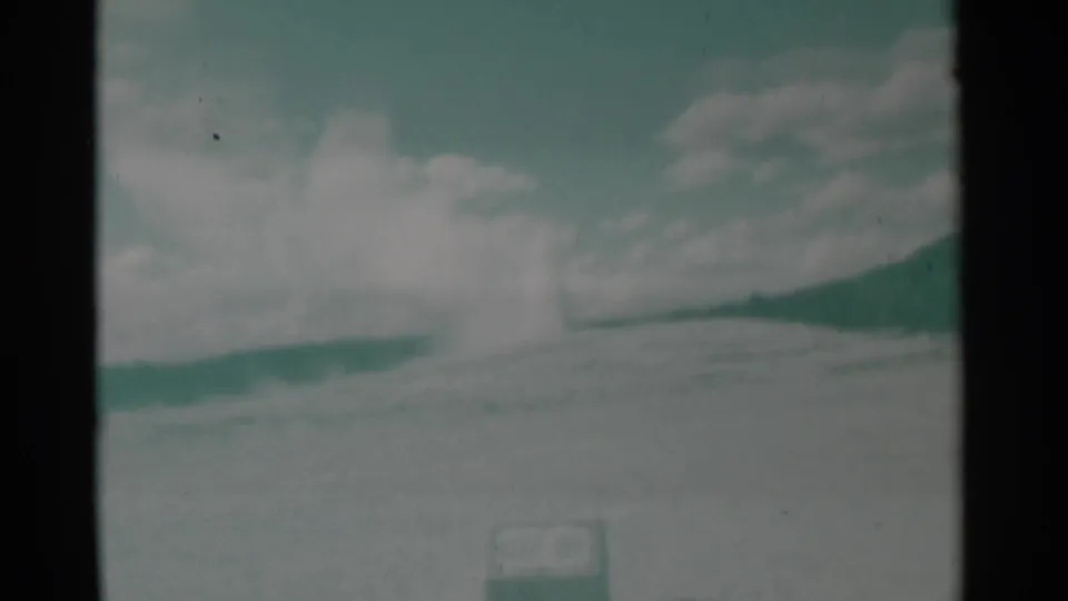 1961 Wyoming home-movie frame with a distant steam plume at a low hill crest, scattered clouds, pale ground, and a small sign near the bottom edge.