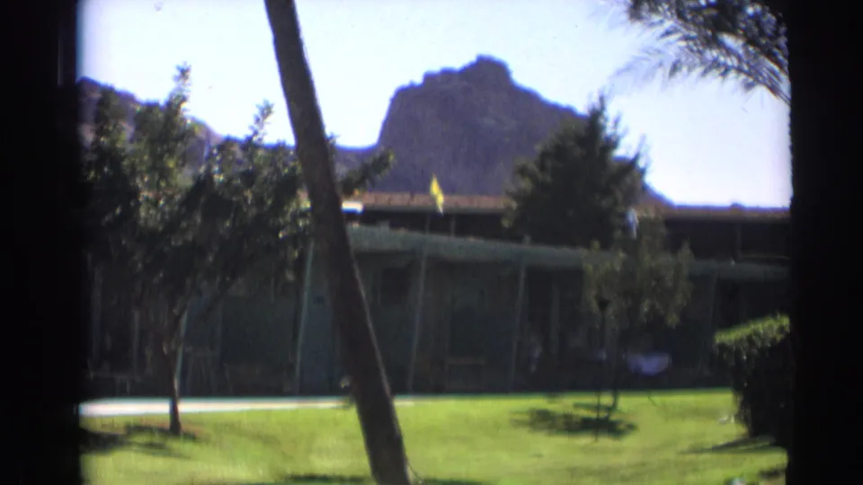1961 8mm frame showing landscaped motel grounds with a palm trunk in front and a rocky mountain rising behind the roofline.