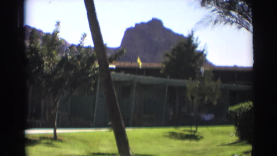 End frame from the 1961 Cheyenne-labeled clip showing motel landscaping, palm trunks, a yellow flag, and a rocky mountain backdrop.