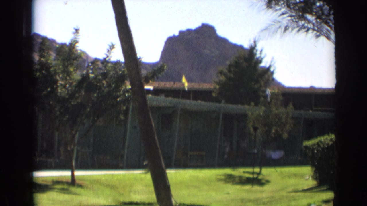 Sunlit lawn and low building framed by trees, with a flag and rocky hill in the background.