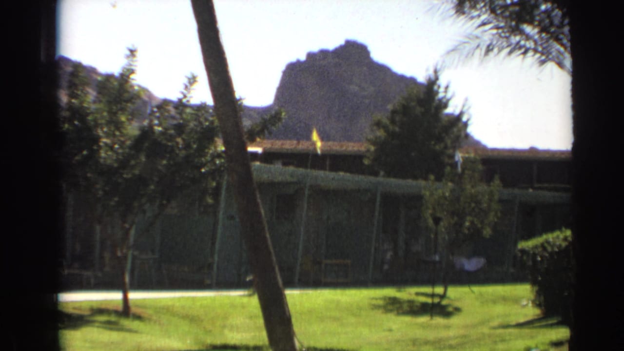 1961 Cheyenne, Wyoming home-movie frame showing a low cabin beyond a green lawn, with trees and a rocky mountain in the background.