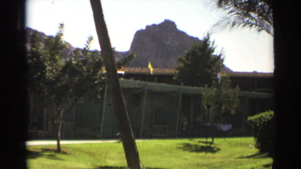 1961 Wyoming home-movie frame showing a quiet cabin exterior across a lawn, with a small flag and rocky mountain backdrop.
