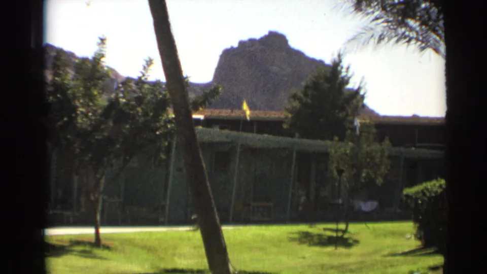 Cheyenne, Wyoming home-movie frame with a low building, lawn, foreground trunk, and rocky butte under bright daylight.