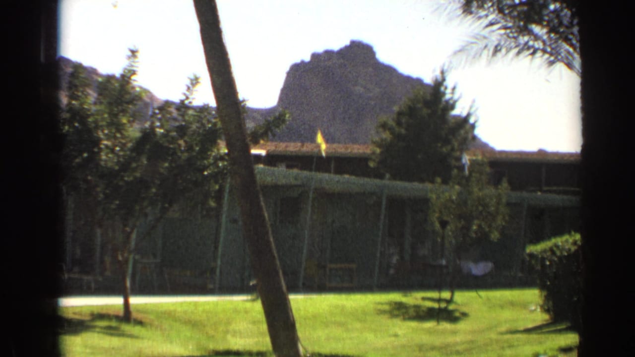 1961 Cheyenne home-movie frame showing a sunlit motel courtyard lawn, small trees, shaded walkway, and rocky hill beyond.