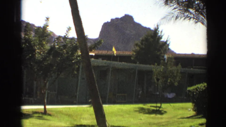 1961 Cheyenne frame holding on the landscaped motel courtyard, shaded low building, palm fronds, and rocky hill under bright sky.