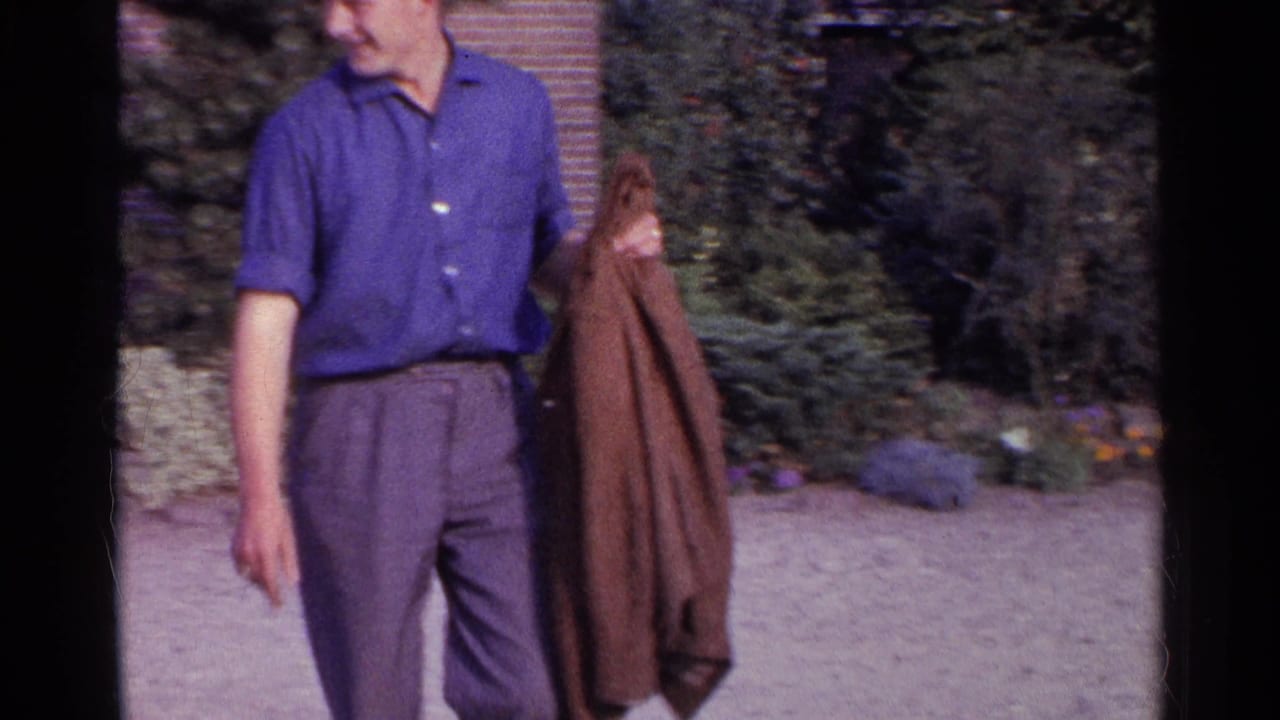 Young man in 1961 Germany standing outdoors by a brick house and greenery while holding a brown jacket.