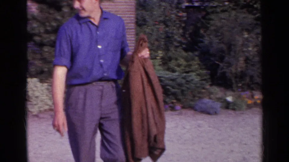 Young man in 1961 Germany standing outdoors by a brick house and greenery while holding a brown jacket.
