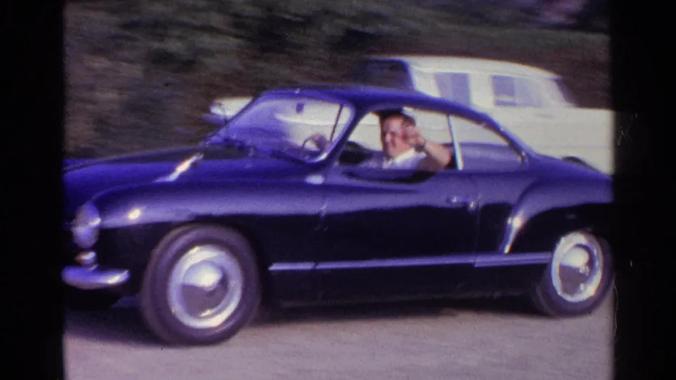 Dark classic coupe on a road in Germany, with a driver visible and waving from inside the car.