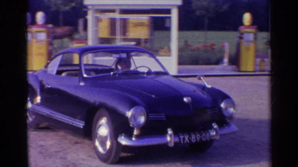 Dark rounded coupe parked at a service-station forecourt in 1961 Germany, with fuel pumps and signage behind.