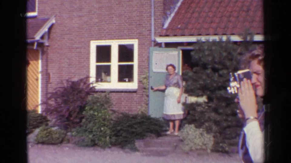 Woman in an apron greets from a brick house doorway in Germany as another person records with a handheld camera.