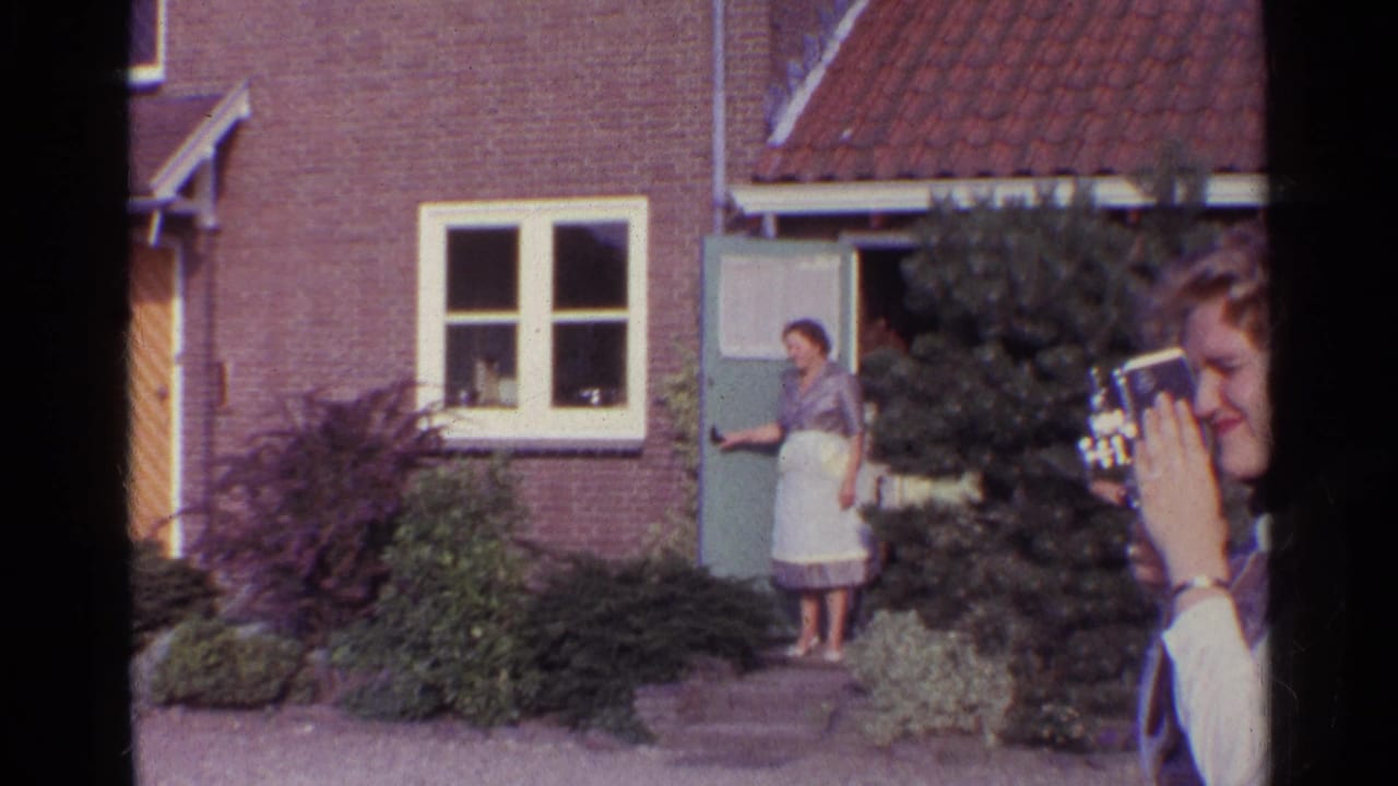 1961 Germany home-movie frame showing a woman at a brick house doorway as a young person holds a small camera in the foreground.