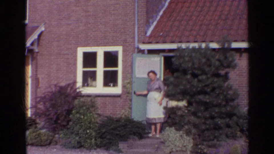 1961 Germany home exterior with a woman in an apron standing in the doorway of a brick house, framed by shrubs and windows.