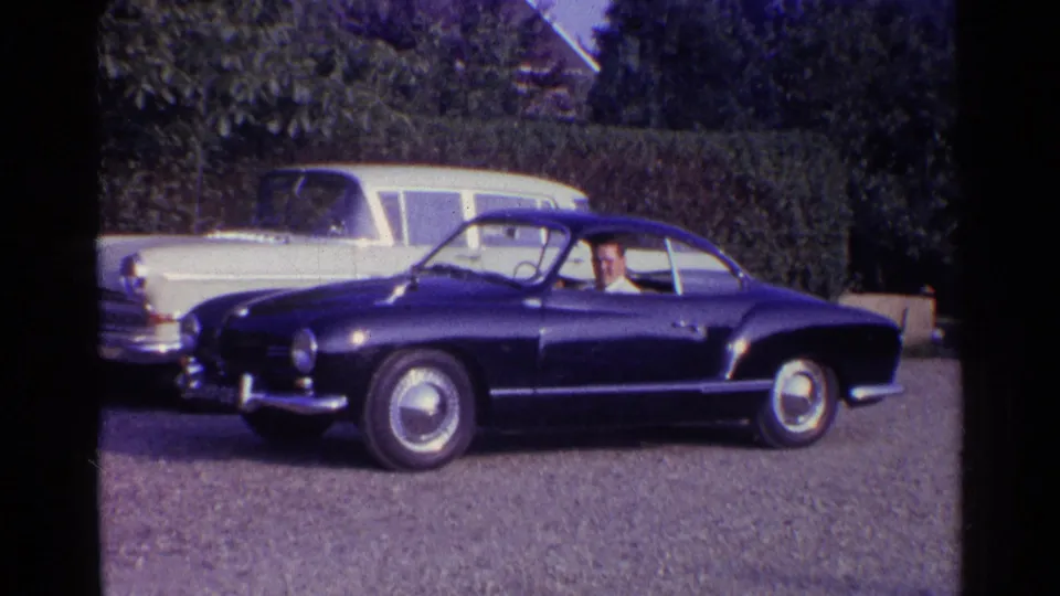 1961 Germany driveway scene with a blue classic coupe, a seated driver, a white station wagon, and hedges behind the cars.