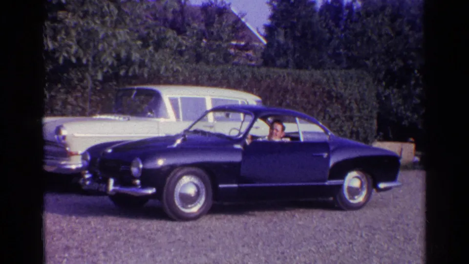 1961 Germany home-movie frame showing a side view of a blue coupe with a driver, parked near a white station wagon and hedge.