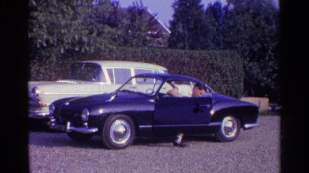1961 home-movie frame showing a deep blue classic coupe in a suburban driveway, with a light station wagon, hedge, and trees behind.