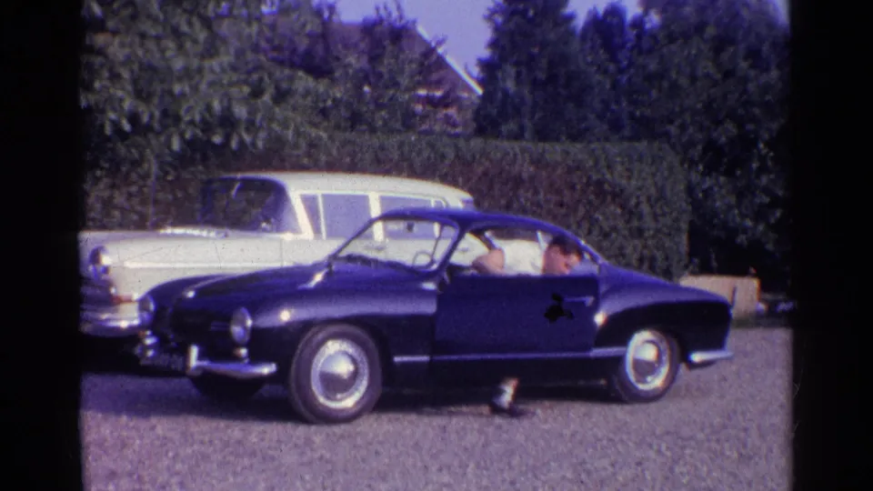 1961 home-movie frame showing a deep blue classic coupe in a suburban driveway, with a light station wagon, hedge, and trees behind.